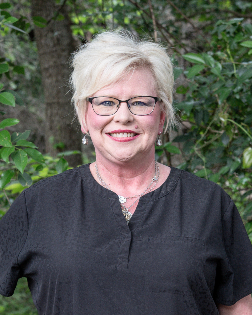 Meet the Team: A woman with short blonde hair, glasses, and a black blouse stands outdoors in front of green foliage, smiling at the camera.