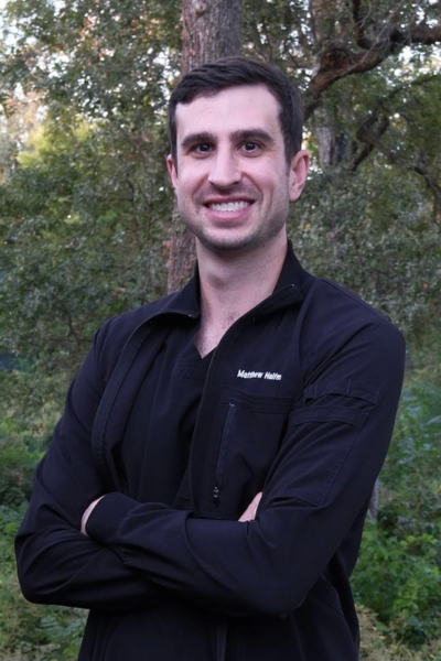 Man with dark hair and a black jacket standing outdoors in Waco with arms crossed and trees in the background, giving the confident look of a local dentist.